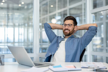 Young man wearing eyeglasses feeling relaxed and smiling, leaning back in his chair with hands behind head, taking a break from working with laptop at modern office