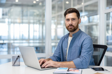 Fototapeta premium Bearded man sitting at a desk with a laptop, looking directly at the viewer in a modern office, representing concepts of remote work, business, technology, and focus