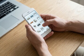 Hands of a person browsing furniture collection on a smartphone while sitting at a wooden table with a laptop, showcasing modern technology and online shopping experience