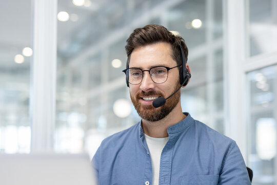 Man wearing glasses and a headset, smiling while looking at a laptop, representing helpful communication and professional customer support in an office setting