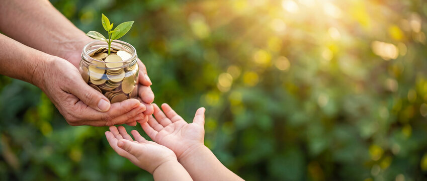 Child&rsquo;s hands receive money jar with coins savings and tiny plant from adult, sunny greenery background with copy space