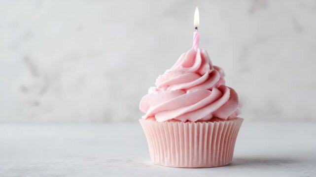 Delicate pink cupcake with swirled icing and lit candle on marble surface