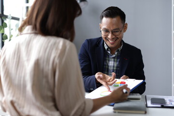 Asian businessman pointing at a financial report with a pen while discussing details with a colleague during a meeting in a corporate office, highlighting teamwork and strategy.