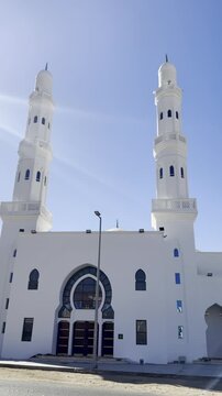 A wide architectural shot of the bright white Masjid Al-Areesh in Medina, showcasing its symmetrical twin minarets and arched entrance under a clear, sun-drenched sky.
