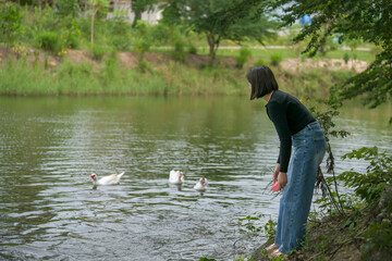 A woman in a black top and jeans stands at the edge of a pond, watching three white ducks swimming peacefully in the water surrounded by nature.