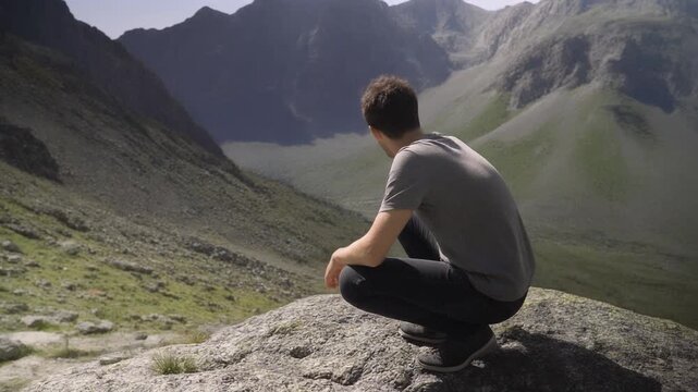 Young man crouching on rock, looking at sunny, remote mountain view 