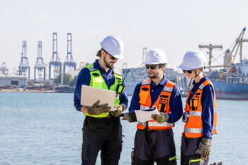 group of industrial engineers workers in a refinery - oil and gas processing equipment and machinery, engineers collaborate with a laptop, blueprint, and digital tablet at the oil storage tanks site.