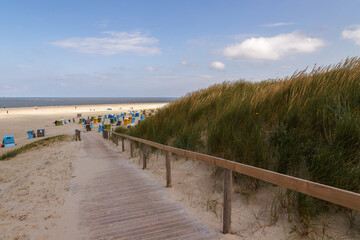 Holzbohlenweg f&uuml;hrt durch die D&uuml;nen mit hohem Gras zu einem Sandstrand mit Strandk&ouml;rben auf der Insel Langeoog