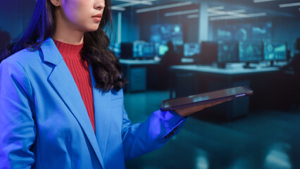 Focused Asian businesswoman analyzes data on digital tablet within high-tech futuristic office. professional dedication in blue-lit cybersecurity or trading command center environment.