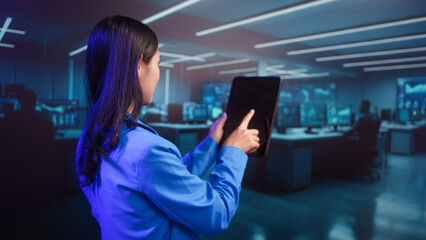 Focused Asian businesswoman analyzes data on digital tablet within high-tech futuristic office. professional dedication in blue-lit cybersecurity or trading command center environment.