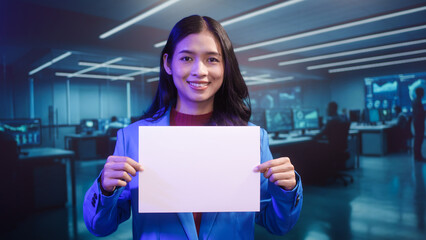 Smiling Asian woman holds blank white paper in high-tech office. This mockup template offers perfect copy space for business messages, advertisements, or announcements in futuristic setting.