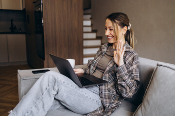 Woman working online from home, using laptop sitting on sofa and drinking coffee