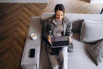 Woman working online from home, using laptop sitting on sofa and drinking coffee