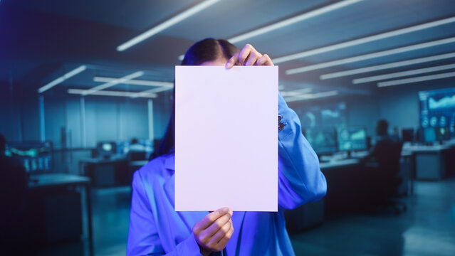 Smiling Asian woman holds blank white paper in high-tech office. This mockup template offers perfect copy space for business messages, advertisements, or announcements in futuristic setting. - Powered by Adobe
