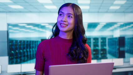Focused Asian female IT professional works on laptop in secure data center. The modern background features server racks, cybersecurity, cloud computing, advanced digital infrastructure management.
