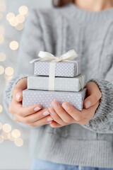 closeup of hands holding wrapped gifts, gray sweater background with white bokeh lights, gift box stack on a woman arms, christmas present concept.