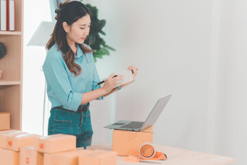 Asian woman ships packages using a laptop. E-commerce order management, logistics arrangements, shipping concept, SME business.