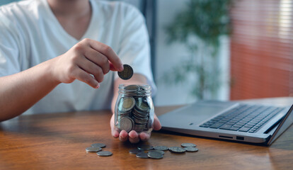 Person putting coins into glass jar beside laptop on wooden table, concept of saving money, personal finance, budgeting, online income, financial planning, investment growth, and future security.