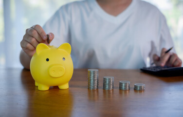 Yellow piggy bank with stacked coins on wooden table, person saving money and calculating budget. Concept of personal finance, savings plan, investment growth, financial discipline at home.