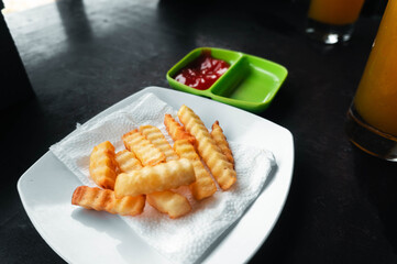 Close-up shot of golden-brown, perfectly-cooked crinkle-cut fries on a pristine white plate, accompanied by a vibrant small bowl of ketchup and refreshing drink.