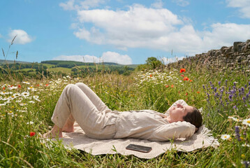 Side view of a relaxed woman wearing linen clothes lying on a woven rug in a field of wildflowers. A smartphone is placed on the mat next to her