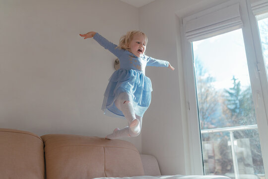 Young girl descends toward sofa wearing layered blue dress, arms open wide, bright daylight enters through window, homemade craft decorations visible across wall, energetic pose captured during indoor