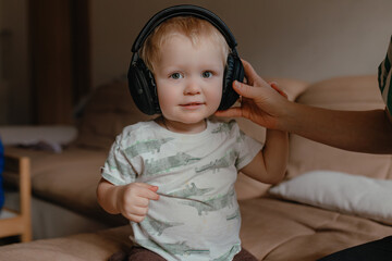 Toddler sitting indoors wearing oversized headphones, woman hand adjusting ear pad, soft daylight over beige sofa, neutral tones, candid relaxed home moment showcasing child interaction with audio dev