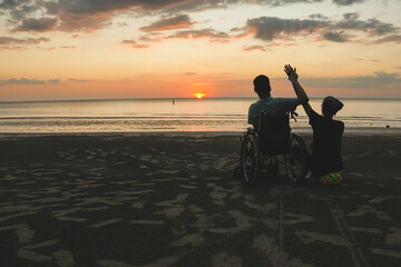 Wheelchair user and family caregiver sitting together on the beach during sunset, enjoying travel and nature, concept of inclusion, accessibility, independence and quality of life.
