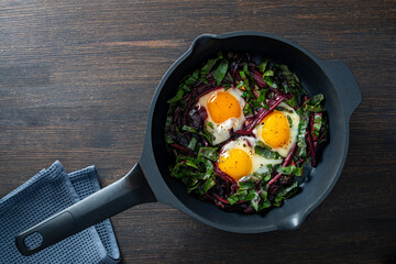 Fried beet stems and eggs in a frying pan on a wooden table, closeup, top view. Food background. Cooked roasted beetroot leaves, eggs with spices for breakfast. Healthy food concept
