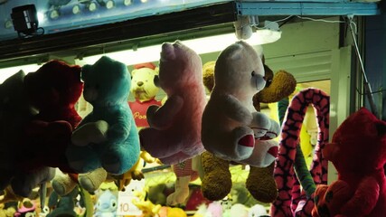 Colorful plush toys hanging as prizes at a fairground game booth. Handheld shot captures festive atmosphere, bright lights and cheerful carnival mood at a festival or amusement park.
