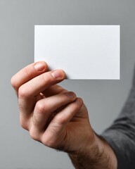 Fototapeta premium A hand holds a blank white card in front of a gray background Concept Minimalist Card Portrait, Hand Holding Card, Blank White Card, Gray Studio Background, Monochrome Mockup