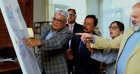 Indian business people discussing projection on chart while senior Asian executives stand close together reviewing growth data on white board inside modern corporate office meeting room - Powered by Adobe