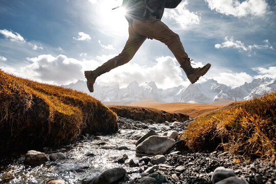 Close up photo of hikers legs jumping over small creek against snow capped mountains with glaciers. Travel active vacations concept