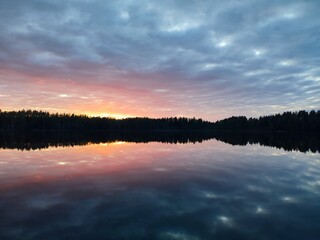 A beautiful view of the sunset, sunrise, against the backdrop of gray, red, orange clouds in the sky, a lake with clear, calm water, and the dark silhouette of the forest on the horizon, late evening