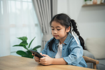 Asian girl using smartphone sitting at a table at home. Cute kid watching the screen, playing...