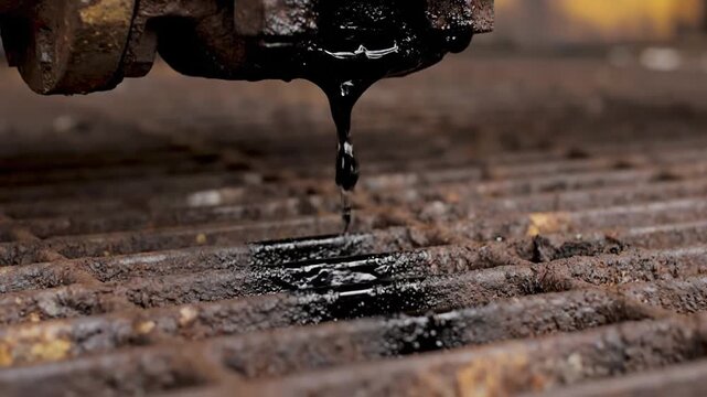 Close-up of black oil leaking and dripping from a rusty industrial pipe onto a metal grate, indicating a dirty environment or machinery malfunction.