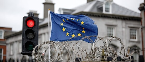 Traffic light shows red with European Union flag above and barbed wire in front of isolated wall with window captured from below with natural lighting