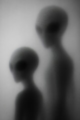 Two abstract alien figures stand side by side. The larger figure is tall and slender, while the smaller one is shorter. Both have large heads and dark eyes.