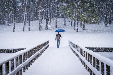 Man walking alone across a bridge with umbrella during winter snowfall. Cold weather, daily commute, loneliness and winter lifestyle concept.