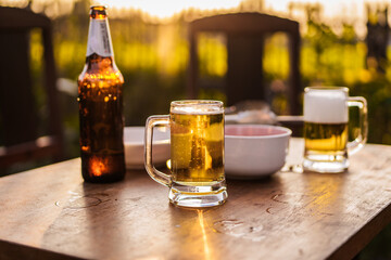 Golden beer in a clear glass mug rests on a wooden table, glowing in warm sunset light outdoor, refreshing beverage moment photography summer relaxation natural bokeh background.