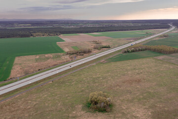 Droga przebiegająca przez r&oacute;wninę pokrytą polami uprawnymi, łąkami, kępami drzew. Jest słoneczny dzień. Zdjęcie wykonano z dużej wysokości z użyciem drona.