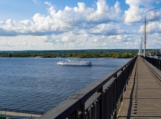 A passenger ship sails along the river. View from above, from the bridge. The walking part of the bridge and the opposite bank of the river with a forest area are visible.