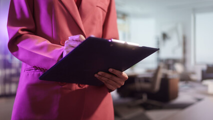 Close-up of a professional businesswoman in a pink blazer analyzing financial statistics on clipboard. She is reviewing data charts and writing notes in a bright, modern corporate office background.