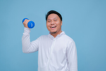 Young Asian man exercising at the gym lifting dumbbells in both hands wearing white t-shirt with happy laughing expression on turquoise blue background 