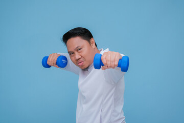 Young Asian man exercising at the gym lifting dumbbells in both hands wearing white t-shirt with happy laughing expression on turquoise blue background 