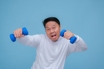 Young Asian man exercising at the gym lifting dumbbells in both hands wearing white t-shirt with happy laughing expression on turquoise blue background 