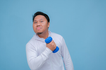 Young Asian man exercising at the gym lifting dumbbells in both hands wearing white t-shirt with happy laughing expression on turquoise blue background 