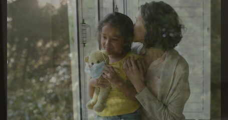 Kissing mother in cardigan cradling child in yellow top at window, with teddy mask and blinds