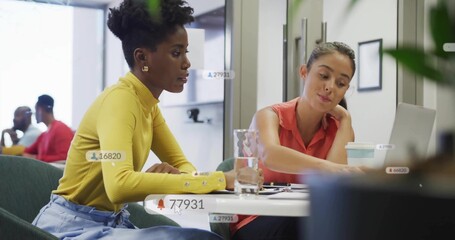 Collaborating pair in yellow and coral tops leaning to laptop at office table with UI labels