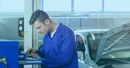 Inspecting mechanic in blue coverall, white tee leaning over tool cart at garage near silver car